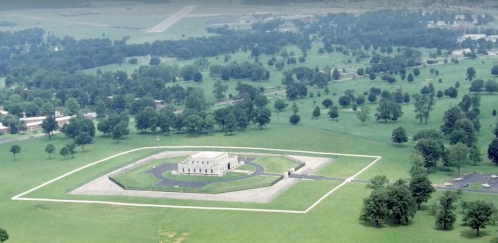 Aerial view of a large, white building surrounded by green fields and trees, with a road leading to it.