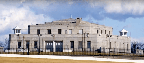 A large, gray stone building with a flat roof and two small towers, set against a blue sky with clouds.