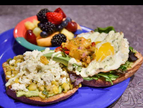 A colorful plate featuring two open-faced sandwiches with toppings and a side of mixed fruit.