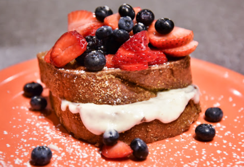 A slice of French toast topped with strawberries, blueberries, and powdered sugar on an orange plate.