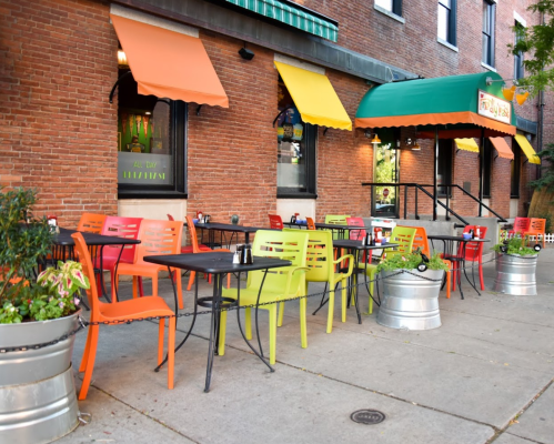 Colorful outdoor seating with tables and chairs in front of a brick restaurant, featuring potted plants and awnings.
