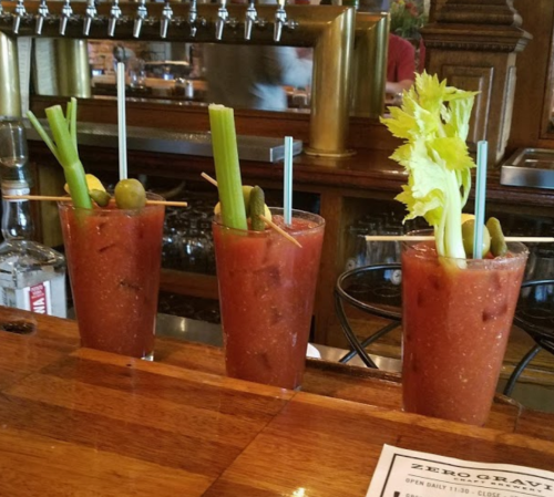 Three Bloody Mary cocktails garnished with celery and olives, sitting on a wooden bar counter.