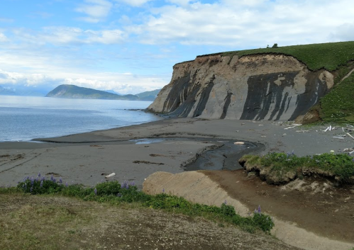 A coastal landscape featuring a steep cliff, sandy beach, and calm waters under a partly cloudy sky.