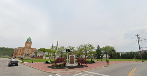 A town square featuring a monument, trees, and a church in the background, with a cloudy sky above.