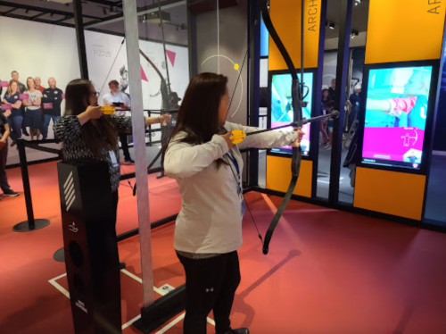 Two women practice archery in a modern indoor setting, with digital displays in the background.