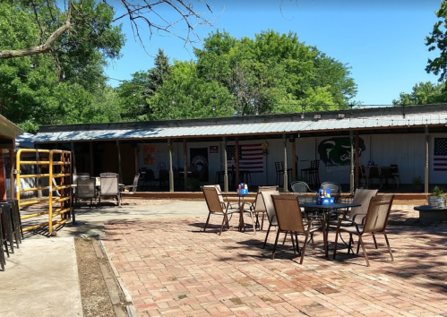 Outdoor seating area with tables and chairs, surrounded by trees and a building featuring flags and murals.