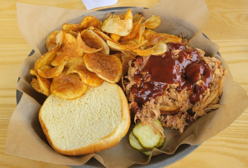 A plate with pulled pork topped with barbecue sauce, served with potato chips, a slice of bread, and pickles.