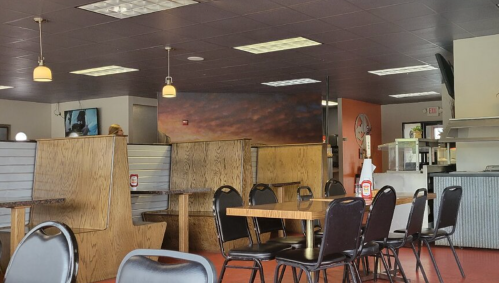 Interior of a casual restaurant featuring wooden booths, black chairs, and a view of the kitchen area.
