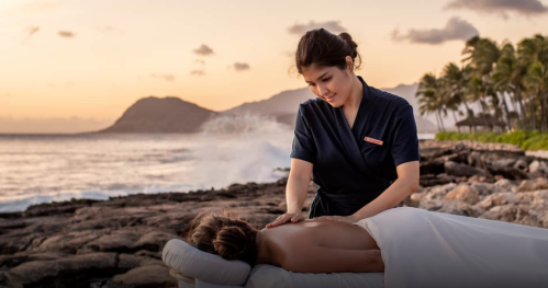 A massage therapist provides a relaxing massage to a client on a beach at sunset, with waves and palm trees in the background.