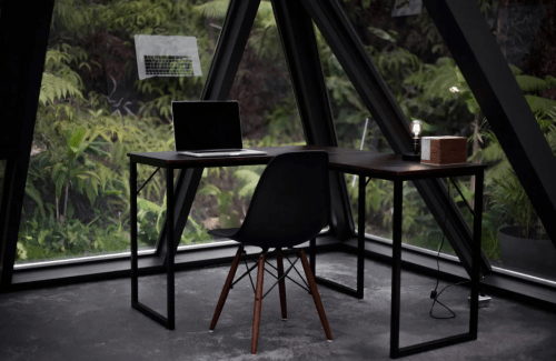 A modern desk with a laptop and a chair, set in a glass-walled room surrounded by greenery.
