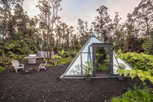 A modern glass pyramid cabin surrounded by lush greenery and black volcanic soil, with outdoor seating and a grill.