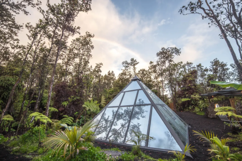 A modern glass pyramid structure surrounded by lush greenery and trees under a cloudy sky.