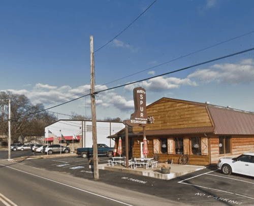 A rustic wooden restaurant with a sign reading "Rusty's Spur" and outdoor seating, set against a clear blue sky.