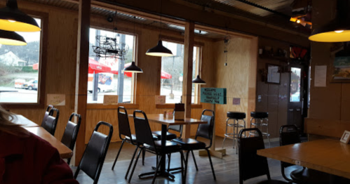 Interior of a cozy restaurant with wooden walls, tables, and chairs, featuring large windows and hanging lights.