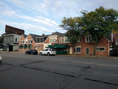 A brick building with green awnings, featuring multiple windows, along a street with parked cars and trees nearby.