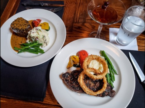 A plate with grilled steak, onion rings, green beans, and mashed potatoes, alongside a plate of beef with vegetables and a cocktail.