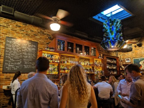 A lively bar scene with a brick wall, shelves of bottles, and a unique chandelier made of bottles.