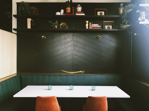 A cozy dining area with a white table, two orange chairs, and a dark wooden wall adorned with plants and decor.