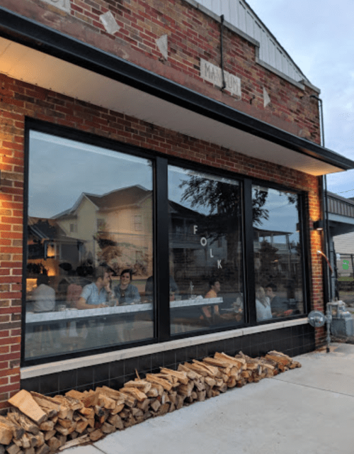 A cozy restaurant with large windows, showcasing diners inside and a stack of firewood outside.