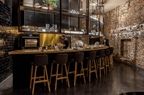 A modern restaurant interior with a bar, high stools, and exposed brick walls, featuring chefs preparing food.