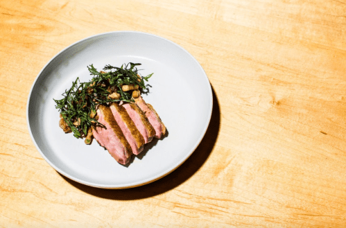 A plate with sliced duck breast served with sautéed greens on a wooden table.