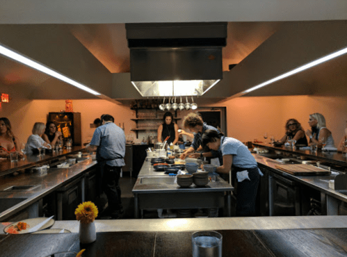 A modern kitchen with chefs preparing food, surrounded by diners at a communal table, warm lighting, and a cozy atmosphere.