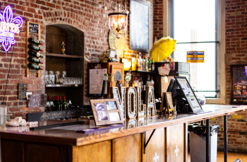 A rustic bar with a wooden counter, brick walls, and decorative lights, featuring a "VOLA" sign and various bottles.
