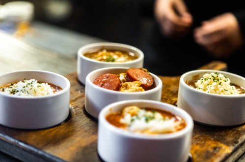 Five small bowls of different stews, topped with cheese and herbs, arranged on a wooden tray.