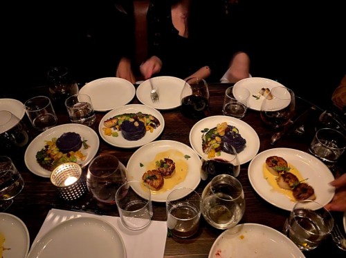 A dimly lit table with several plates of gourmet food, glasses of wine, and hands reaching for dishes.