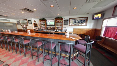 A cozy bar interior with a wooden counter, high stools, and shelves stocked with various drinks. TVs are mounted on the walls.