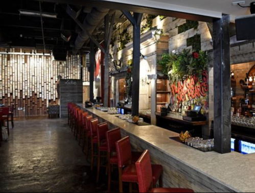 A stylish bar interior with red chairs, a stone counter, and greenery on the walls, creating a warm, inviting atmosphere.