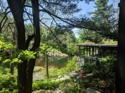 A serene view of a pond surrounded by trees, with a modern glass structure nearby and people enjoying the scenery.