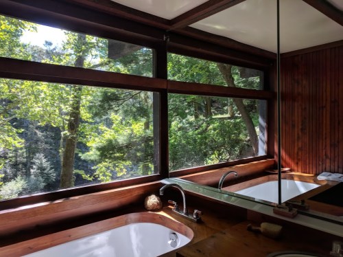 A modern bathroom with large windows overlooking a lush forest, featuring wooden accents and a sleek bathtub.