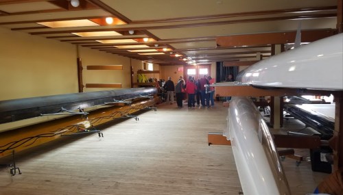Interior of a boathouse with rowing shells on display and a group of people gathered in the background.
