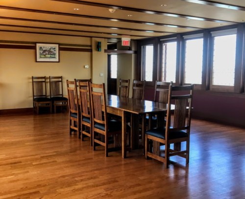 A spacious room with a long wooden table and eight wooden chairs, featuring large windows and wooden flooring.