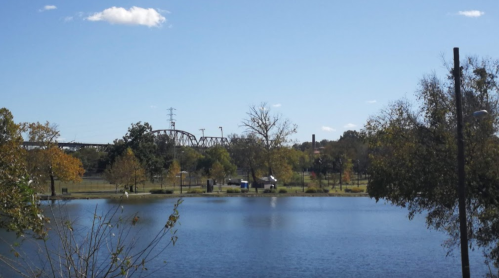A serene lake reflects a clear blue sky, with trees and a distant bridge in the background.