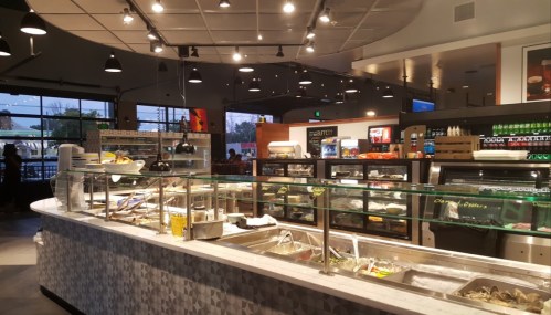 A modern restaurant interior featuring a food display counter, shelves of drinks, and large windows with outdoor views.