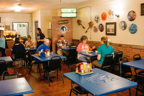 A cozy diner with blue tables, patrons enjoying meals, and colorful wall decor in a warm, inviting atmosphere.