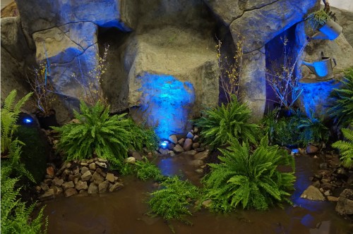 A rocky landscape with blue lighting, ferns, and a small stream flowing through the greenery.