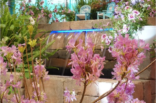 A serene garden scene featuring a small waterfall surrounded by pink flowers and lush greenery.