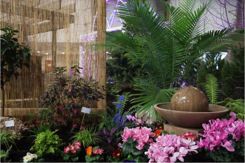 A lush garden scene featuring vibrant flowers, greenery, and a decorative water fountain surrounded by bamboo.