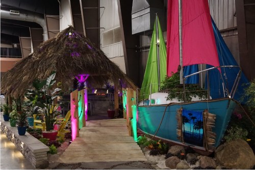 A vibrant tropical-themed setup featuring a thatched hut, colorful sailboat, and lush plants with decorative lighting.