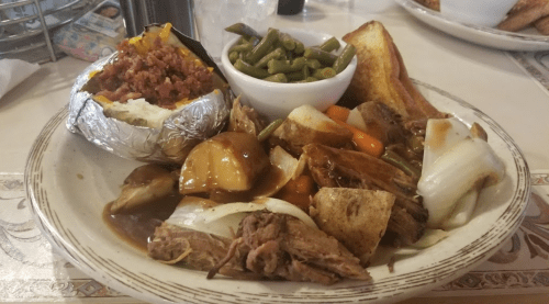 A plate of hearty food featuring pulled pork, vegetables, a baked potato, green beans, and toasted bread.