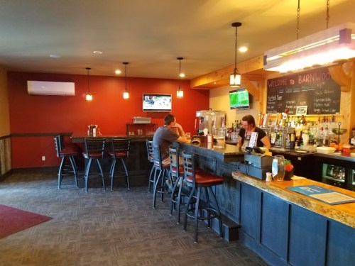 A cozy bar interior with a wooden counter, stools, and a bartender serving drinks. TVs display sports in the background.