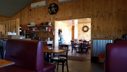 Interior of a rustic restaurant with wooden walls, tables, and chairs, featuring a doorway to another dining area.