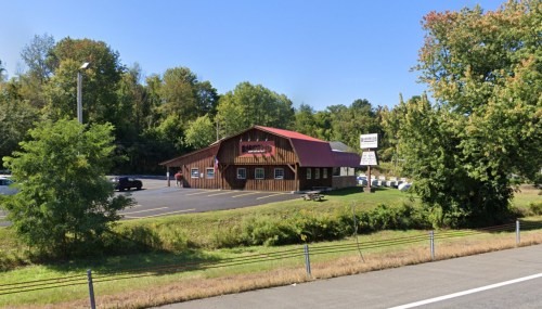 A rustic building with a red roof, surrounded by trees, and a parking lot along a highway.