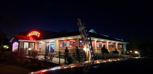 A restaurant exterior at night, featuring bright signage and a line of people waiting outside.