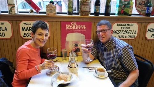 A couple enjoys a meal at a restaurant, each holding a large glass of red wine, with a cozy atmosphere and vintage decor.