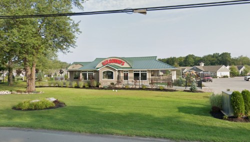 A restaurant with a green roof and stone exterior, surrounded by grass and trees, with parking in the background.