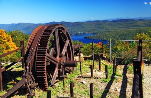 Rusty gear and machinery remnants overlook a scenic lake surrounded by mountains and colorful autumn foliage.